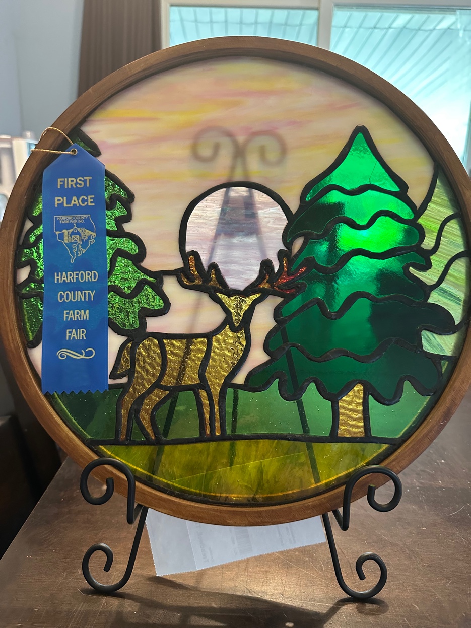 Award-winning stained glass roundel of a deer among pine trees at sunset, with First Place ribbon